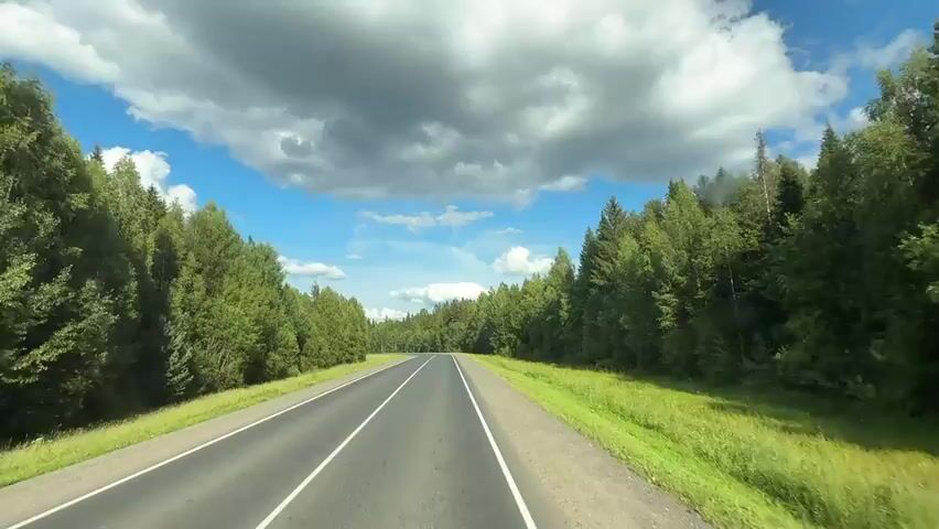 This media file captures a scenic drive on a paved road through a dense, green forest. The road curves gently to the left, with distinct white lines marking the lanes. Lush green grass lines the sides of the road, contrasting with the dark asphalt. The forest consists primarily of tall, coniferous trees, with some deciduous trees interspersed, creating a vibrant and varied green landscape.

The sky above is a striking blue, dotted with large, dramatic white clouds that cast shadows on the scene below. The lighting suggests it is daytime, likely mid-morning or afternoon, given the brightness and the way the clouds are illuminated. There are no people or man-made objects visible other than the road itself and what appears to be a dashed white line to the right, possibly indicating the edge of the road or a shoulder.

The overall mood is peaceful and expansive, evoking a sense of journey and immersion in nature. The location is identified as Volokovye, Russia, which aligns with the characteristic boreal forest landscape depicted. No text is visible in the media.
