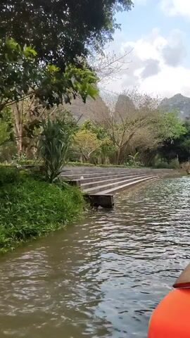 The video depicts a scene from a boat moving on a body of water surrounded by lush greenery and tall, rocky mountains. The water appears calm with gentle ripples.  On the left bank are concrete steps leading down to the water's edge, framed by vegetation. As the video progresses, the camera angle shifts to reveal a person wearing a traditional conical Asian hat and an orange life vest.  Other small boats can be seen in the distance, each carrying people who appear to be enjoying the serene surroundings. The sky above is partly cloudy. The overall impression is one of natural beauty and peaceful exploration.