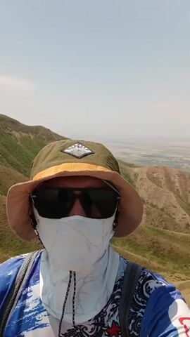 A first-person perspective video shows a person wearing a bucket hat, sunglasses, and a face mask. They are standing on a grassy, hilly landscape. The person is wearing a blue and white shirt with a pattern and a backpack. The background shows rolling hills and a hazy sky. The camera pans slightly, revealing a path winding through the hills.