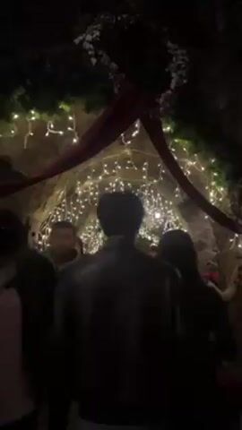The video shows a group of people walking through a stone arched tunnel decorated with strings of white lights. The tunnel is low and the group walks slowly. The lighting is dim, with the white string lights providing the main source of illumination. Decorations are visible along the top edges of the tunnel. People in the group are wearing casual clothes.