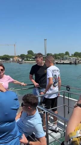 The video shows a group of people on a boat ride, likely on a sunny day. The boat is moving through the water, leaving a wake behind it. Several individuals are visible, including two young men standing near the railing, a woman in a pink shirt who appears to be taking pictures with her phone, and another person in a blue shirt sitting down. A construction crane is visible in the background on the shoreline. The overall atmosphere seems relaxed and touristy.