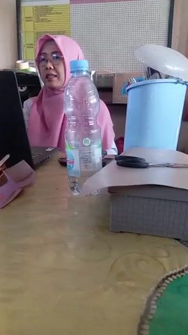 A woman wearing a pink hijab and glasses is sitting at a table. In front of her is a laptop and a plastic bottle of water. There are also scissors and a box on the table. Behind her, a blue trash can is visible, and a whiteboard with writing on it hangs on the wall.