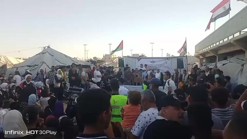 This video captures a gathering of a large crowd of people in Nuseirat, Palestinian Territories. The scene appears to be set outdoors, possibly in a refugee camp or a community space, with several large tents visible in the background. The time of day is late afternoon or early evening, indicated by the warm, golden light and long shadows. The sky is clear, suggesting fair weather.

The crowd is diverse, with men, women, and children present. Many of the women and girls are wearing hijabs. People are densely packed, facing towards a raised platform or stage area where several individuals are standing. A banner with Arabic text is displayed behind them. Two flags are visible: one with the Palestinian flag's colors and another appearing to be the Iraqi flag. A person in a red academic-style robe is standing in the foreground among the seated audience.

The atmosphere seems to be one of a public event or gathering, possibly a rally, a community meeting, or a cultural performance. The expressions on people's faces are varied, but many appear engaged and attentive. Some individuals in the crowd are holding up their phones, suggesting they are recording the event. The presence of tents and the large gathering of people might indicate a response to a humanitarian situation or a significant community occasion. The camera's watermark "Infinix HOT 30Play" is visible in the bottom left corner of the frame.