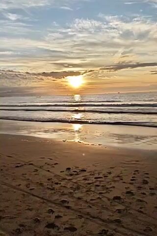 A serene ocean sunset is captured in this vertical video. The sun, a bright orb, is positioned slightly left of the center of the frame, casting a golden reflection on the calm water. The sky above is a mix of blue and soft orange and yellow hues, streaked with wispy clouds that are catching the sunset light. Gentle waves are rolling onto a sandy beach in the foreground. The sand itself bears faint tire tracks and scattered footprints, indicating recent activity. The overall atmosphere is peaceful and tranquil, highlighting the natural beauty of the coastal landscape at dusk.