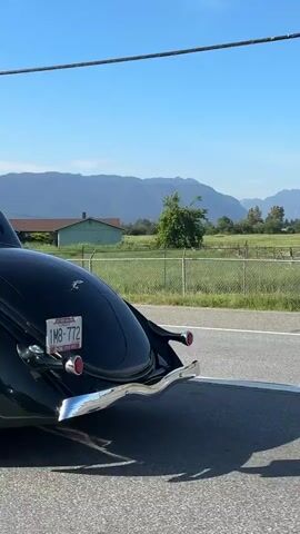 The video shows a dark green vintage coupe driving on a paved road. The car has a chrome bumper and running boards and shiny modern wheels. Mountains are visible in the background under a clear blue sky. The car has a license plate that reads "1MB 772". The video is shot from another car driving alongside the coupe.