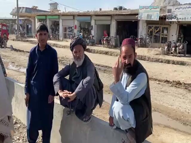 Three men are posed outdoors in front of a row of buildings and shops. The youngest, on the left, is standing with arms at his sides and wearing a dark, long-sleeved garment. The man in the center is squatting on top of a concrete structure with hands clasped and wearing a dark turban and dark, long-sleeved garment. The man on the right is squatting on the same concrete structure with his left hand under his chin and wearing a light blue, long-sleeved garment and a dark vest. The area behind them has small shops and motorcycles parked. A muddy road is visible in front of the men. The sky is clear and the light suggests it is daytime.