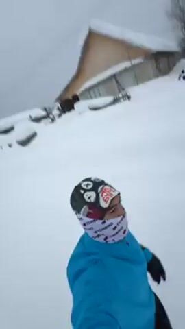 The video begins with a first-person perspective of a person sledding down a snow-covered hill. They are wearing a blue jacket, black gloves, a helmet, and a face covering. In the background, there is a building partially covered in snow. The camera angle is slightly tilted as the person slides downhill. 

The video then cuts to a scene of three people walking down a snow-covered road. The person in the middle is wearing a red jacket, black pants, and a hat. The other two people are partially visible on either side of them.  The environment consists of snow-covered ground, houses, and trees. The sky is overcast.