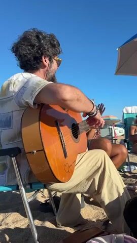 A group of people are gathered on a sandy beach on a sunny day. In the foreground, a man with curly dark hair and a beard is playing an acoustic guitar. He is wearing a patterned short-sleeved shirt and light-colored pants. He has several bracelets on his left wrist and tattoos on his arm. He is sitting in a folding chair. To his right, another person, whose face is obscured by sunglasses, is also seated and clapping their hands. Further to the right, two women are seated, also clapping their hands. One woman is wearing a white bikini top and sunglasses, and the other is wearing a colorful patterned swimsuit. Beach umbrellas and chairs are visible in the background, along with a cooler and bags. The ocean and a clear blue sky are visible behind the people. The overall scene suggests a casual, festive gathering on the beach.