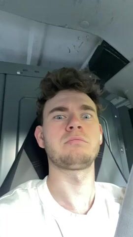 A young man with curly brown hair and blue eyes is in what appears to be the passenger seat of a vehicle. He is wearing a white t-shirt and white wireless earbuds. His facial expression shifts between wide-eyed surprise and a more neutral, pensive look. The background shows the interior of the vehicle, including a gray panel and a seatbelt. The lighting is somewhat dim, suggesting an indoor or possibly nighttime setting.