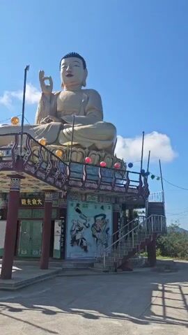 The video shows a large golden Buddha statue. The camera pans from the Buddha statue to a row of colorful lanterns hanging on a wire. The lanterns are red, orange, yellow, green, blue, and pink. The video then pans to a landscape with trees and hills.