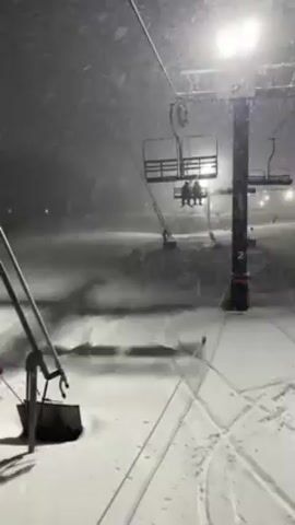 The video shows a snowy scene at a ski resort at night. A ski lift is visible, with a couple riding uphill on one of the chairs. Snow is falling heavily, and the ski slope is illuminated by overhead lights. Ski tracks can be seen in the snow. The video seems to be taken from a stationary position near the base of the ski lift.