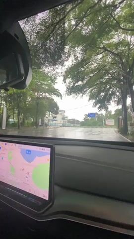 The video is shot from inside a car, looking through the windshield. It is raining, and the windshield wipers are intermittently clearing the view. The road ahead is wet and reflects the overcast sky. Tall trees with green foliage line both sides of the road. In the distance, there are some buildings and a blue sign with white text. The car's dashboard is visible in the foreground, with a large touchscreen display showing a navigation map. The overall scene suggests a drive on a rainy day through a suburban or semi-rural area.