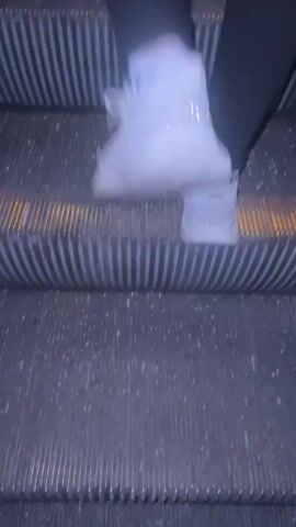 The video shows a close-up view of a person's legs and feet as they walk up an escalator. The person is wearing white sneakers with some sparkle embellishments and black leggings. The escalator steps are metallic and have a ribbed texture with yellow safety lines. The lighting is dim, with some warm yellow lights visible from the escalator's mechanism. The camera angle is low, focusing on the movement of the feet on the steps.