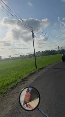 The video begins with a close-up view of a motorcycle's side mirror, reflecting a person's hand and arm. The perspective then shifts to a first-person view from a moving vehicle, likely a motorcycle, traveling along a paved road. To the left of the road is a lush green rice field, and to the right, a smaller paved shoulder. Overhead, a network of telephone or power lines crisscrosses the sky, supported by utility poles. In the distance, more utility poles and trees are visible, as well as a hazy mountainous or hilly landscape. The sky is blue with scattered white clouds. Several motorcycles are seen on the road ahead. One motorcycle is traveling in the same direction as the viewer and is in the foreground. Another motorcycle is further ahead and to the left of the viewer. The scene suggests a rural or semi-rural setting.