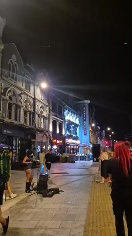 At night on a commercial street, two women busk with a microphone and guitar amp. There are several pedestrians walking past and people gathered in small groups nearby. The buildings lining the street are multi-story and have illuminated signs.