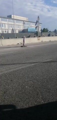 The video shows a view from a moving vehicle on a highway. The road surface is asphalt with white lines marking the lanes. On the left side of the road, there is a concrete barrier, a tall pole with a traffic sign, and a building. Further down the road, there are trees and a cloudy sky. On the right side of the road, there are more concrete barriers with red and white striped markers, and what appears to be a large sign or structure. A white car is visible in the distance on the right side of the road. The overall environment appears to be an urban or suburban area during the daytime.