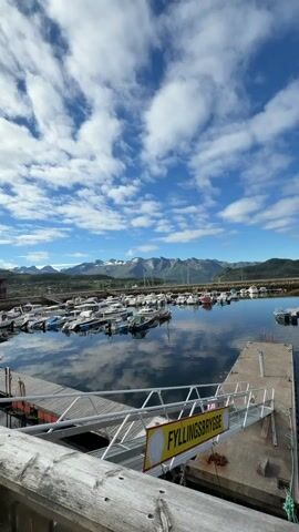 This vertical video captures a serene marina scene in Ørnes, Norway. The main subjects are a fleet of white motorboats docked in calm, reflective water under a bright blue sky dotted with fluffy white clouds. In the foreground, a weathered wooden railing partially frames the view, with a metal ramp and a concrete pier extending into the water. A sign with yellow text reads "FYLLINGSBryGGE" (filling quay).

The setting is a picturesque coastal area, with a backdrop of rolling green hills and distant, snow-capped mountains. The sun is high in the sky, indicating it is daytime, likely late morning or early afternoon, given the clear weather and bright sunlight. The overall mood is peaceful and tranquil, with the gentle ripples on the water and the stillness of the boats suggesting a quiet day. The presence of boats indicates a maritime community or a popular spot for recreational boating.