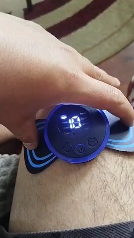 A close-up shot shows a person's hand pressing down on a round, blue-lit device attached to their knee. The device has a digital display showing the number "10" and several buttons. The device appears to be a muscle stimulator or massager. The background is blurred, showing a carpeted floor with a patterned rug.