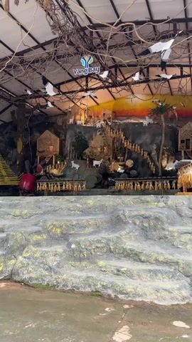 This video shows a staged scene depicting a village or farm setting. In the foreground, there is a rocky, textured surface. The background features a man-made rock formation with several small, rustic-looking buildings. White cutouts of goats are placed around the rock formation, some on ledges and some appearing to climb stairs. Garlands of what look like dried corn hang along horizontal poles in front of the rock face. At the top of the scene, decorative white birds, possibly cranes, are suspended from the ceiling or overhead structures. A logo reading "Yang Bay" is visible near the top center of the frame. The overall impression is of a diorama or exhibit designed to represent a rural environment.