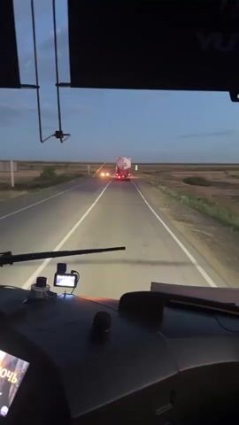 This video captures a forward-facing view from the interior of a vehicle, likely a truck or bus, as it travels along a paved road in Бастау, Kazakhstan. The time of day appears to be dusk or early evening, with a twilight sky overhead transitioning from a soft blue to darker hues. The weather seems clear, with only a few wispy clouds visible.

The main subjects are the road stretching ahead and the surrounding landscape, which consists of flat, open terrain with dry vegetation, characteristic of a steppe environment. A white tanker truck with its red taillights illuminated is visible in the distance, traveling in the same direction. Shortly after the video begins, a white car with its headlights on approaches from the opposite direction and passes the vehicle.

Visible inside the cabin are parts of the dashboard, including a GPS device or camera mounted on the windshield, a windshield wiper, and a screen displaying text in Russian. The visible text on the screen appears to be the title of a song or album: "Ночь" (Noch', meaning "Night") and "cover" (possibly indicating a cover version).

The overall mood conveyed is one of travel and the quiet solitude of a journey during twilight. The lighting from the vehicle's headlights creates illuminated streaks on the road, highlighting the asphalt and the white dividing lines. The scene is peaceful and the sense of movement is constant, suggesting a long-distance drive.