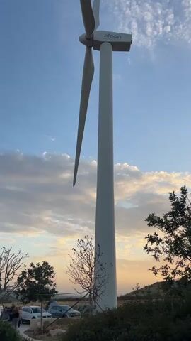 The video shows a large, white wind turbine on a clear day with a sky partially covered by light clouds. The wind turbine has the brand name "Afcon" on its nacelle. The blades are still, not rotating. In the background, a landscape of low hills and trees is visible. There is also a parking area where several cars are parked. A group of people are walking along a pathway near the base of the turbine. The sky has hues of blue and hints of orange, likely from a sunset or sunrise.