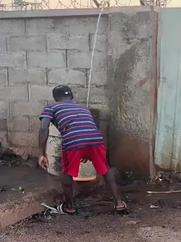 The video shows a man filling a bucket with water from a pipe. He is wearing a striped shirt, red shorts, and sandals, and a baseball cap. He places the bucket under the pipe, and when it's full, he moves it. The area around the pipe is wet and muddy. There is a brick wall behind him and a fence with barbed wire on top. A metal barrel stands next to the wall.