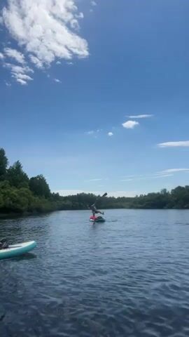 A person on a paddleboard performs a handstand in the middle of a lake, holding a paddle. Another paddleboard is visible to the left, partially in frame. Further in the distance, two individuals are in the water, one appearing to be swimming towards a floating object or raft. The lake is surrounded by lush green trees and vegetation under a bright blue sky with scattered white clouds. The water is calm with a few ripples.