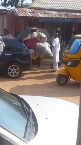 A group of people are gathered around the back of a black hatchback car, which is heavily loaded with various items strapped to the roof. The scene is outdoors on a dirt road, with buildings in the background, including a shop with a corrugated iron roof. A yellow auto rickshaw is parked nearby. Some people are standing around the car, while others appear to be loading or securing the items. One person is wearing a yellow uniform. The scene is viewed from inside another vehicle, as evidenced by the visible windshield and hood.