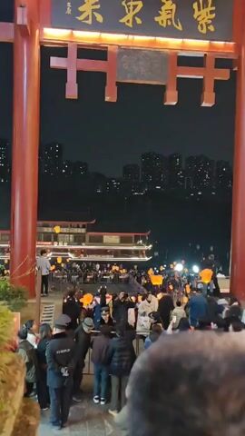 The video shows a nighttime scene in a Chinese city with shops and restaurants. The camera begins facing a large red archway, and then it turns to show a man standing on the steps in front of a restaurant called "LAOKAI" with signs in Chinese. The camera then pans up to show a steep staircase lined with buildings on either side. People are walking up and down the stairs. Some of the buildings are decorated with red lanterns. Trees overhang the staircase.