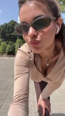 A slender woman wearing sunglasses and athletic clothes stretches and dances on a paved urban plaza on a sunny day. The woman is wearing a long-sleeved beige top, brown flare yoga pants, and white sneakers. She has brown hair pulled into a ponytail. She stretches briefly and then breaks into a short dance, moving her arms and legs in a rhythmic fashion. At one point she poses with her hand on her rear. In the background are tall buildings, green trees, and a white vehicle passing by.