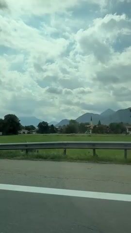 The video shows a landscape viewed from a moving vehicle. In the foreground, there is a road with a white line. The middle ground consists of a green field with some posts. In the background, there are trees, buildings that appear to be a small village or town, and mountains under a cloudy sky. There are no people visible. The video is very short, lasting only a few seconds.