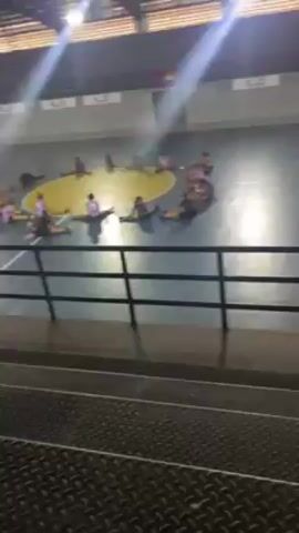 The video shows a group of people stretching in a circle on a basketball court. The court is indoors in a large gymnasium-like space. There are black railings in the foreground. The lighting is bright. The people are wearing athletic clothes. They are sitting on the floor of the court, stretching.