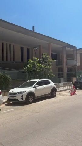 This media file captures a view of a street in Riudoms, Spain, on a clear, sunny day. The camera pans from left to right, showcasing parked cars, a building, and a pedestrian walkway.

In the foreground, two cars are visible on the street. A white SUV is parked on the left, and a dark SUV is parked further down on the right. The dark SUV has a license plate that appears to read "4309 LCS". Orange traffic cones are placed near the dark SUV, suggesting some form of roadwork or obstruction.

The background features a building with a modern architectural style, characterized by its flat roof and balconies. Adjacent to this building, residential structures with balconies and windows are lined up along the street. A pedestrian crossing is marked on the road with red and white stripes, and a pedestrian crossing sign is prominently displayed on a lamppost on the right side of the street. The sky is a bright, cloudless blue, indicating fair weather and likely midday. The presence of streetlights and the well-maintained pavement of the sidewalk suggest an urban or suburban environment. No people are visible in the frame. The overall impression is a quiet, ordinary street scene on a pleasant day.