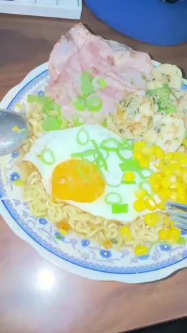 The video shows a close-up, top-down view of a plate of food. The plate is round with a blue and white floral pattern around the rim. The food consists of instant ramen noodles, a fried egg with a bright orange yolk in the center, corn kernels, several pieces of cooked shrimp, and slices of ham or bacon. The dish is garnished with chopped green onions. A fork is visible on the right side of the plate, and a spoon is on the left. The food is placed on a wooden surface. The lighting suggests an indoor setting, possibly a table. The camera is stationary for the entire clip.