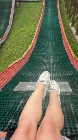 A first-person perspective video shows someone sitting on a green mesh slide with red and yellow borders. The slide appears to be part of an outdoor amusement or activity, with wooden structures and grassy areas visible in the background. The person is wearing white sneakers and white socks, with their legs and feet positioned at the top of the slide, ready to descend. The camera angle provides a clear view of the slide ahead and the surrounding environment. The video is very short, capturing only a few seconds of the experience.