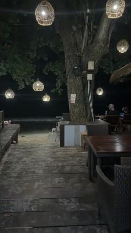 The video captures a night scene at a beachfront restaurant or cafe. The shot is stable, showing a wooden walkway or steps leading down to a sandy beach. Overhead, several spherical, woven-style light fixtures hang from trees, casting a warm glow. The ocean is visible in the background, with waves breaking on the shore under the dark night sky. On the right side of the frame, parts of tables and chairs are visible, suggesting outdoor dining. A large tree trunk stands prominently on the right, decorated with some lights and a white sign with a red circle and a diagonal line, possibly indicating a restriction. People are faintly visible sitting at tables further down the beach and near the water, enjoying the ambiance. The sounds of crashing waves are audible throughout the video, creating a serene atmosphere.