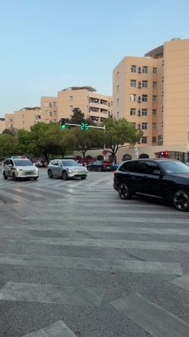 The video shows a medium shot of an urban intersection. In the foreground is a gray crosswalk, its gray lines visible. A white police vehicle with blue and red flashing lights on its roof is traveling across the crosswalk from left to right. To the right of the police vehicle, a silver SUV is also crossing the crosswalk. Both vehicles are moving, and the traffic light above them is green. In the background are several tan apartment buildings, arranged in a row. Between the street and the buildings are trees. People can be seen standing near the crosswalk and on the sidewalks near the buildings. A black car passes on the right side of the frame. The sky is a pale blue, suggesting either early morning or late afternoon.