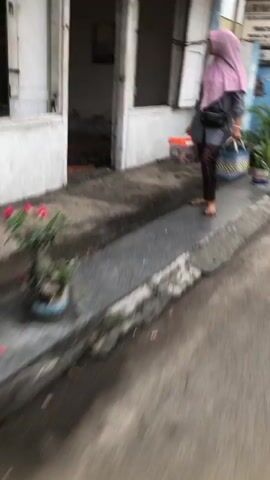 The video is extremely blurry, making detailed analysis difficult. A person wearing a pink hijab and dark patterned leggings is walking away from the camera on a paved walkway. They are carrying a dark bag over their shoulder and holding a blue woven basket in their right hand. To the left of the walkway is a small potted plant with red flowers. Behind the walkway is a building with white siding and open doorways. The ground appears wet in some areas, suggesting it may have recently rained.