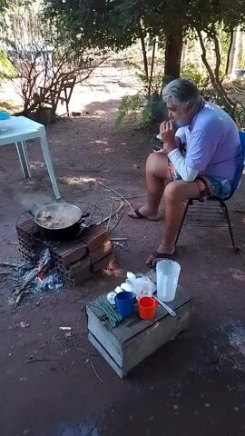 A man with gray hair, wearing a light purple t-shirt and patterned shorts, sits in a blue chair outdoors. He is looking down and appears to be holding something near his mouth. In front of him, a pot of food is cooking over an open fire built with bricks. To the right of the pot, on a wooden crate, are two cups (one blue, one orange), some green items that look like herbs, a white paper packet, and a clear plastic cup. The ground is dirt, and trees and foliage are visible in the background. A white plastic table is to the left of the frame.