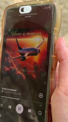 A hand holds a smartphone displaying music playing on a music app. The song is titled "Volver a empezar" by Snag1795 and Miriam Disafi. The album art features an airplane flying into a sunset. The background is out of focus.