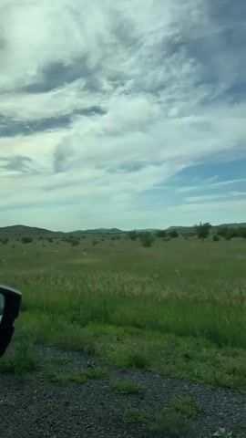 A car drives down a gravel road, passing a vast field of green grass. The sky is blue with scattered white clouds. In the distance, rolling hills are visible. A black car mirror is visible on the left side of the frame.