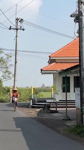 A person wearing a conical hat and an orange shirt rides a bicycle down a paved road. The person is moving from left to right in the frame. To the left of the road, there is a utility pole with wires. Behind the utility pole, there are trees and vegetation. To the right of the road, there is a white building with a red tiled roof and a green awning. The building appears to be some kind of structure, possibly a place of worship or a municipal building. There are stairs leading up to the building. The sky is clear and blue. The ground is dirt and asphalt. In the distance, there is a reddish-brown structure, possibly another building or a monument.