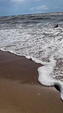 The video shows a beach scene with waves crashing on the shore. The sand is wet from the receding tide, and footprints are visible. In the distance, several people are seen wading in the water and walking along the beach. The sky is cloudy with patches of blue visible. The waves are choppy, suggesting a windy day. The overall atmosphere is one of a lively beach on a somewhat overcast day.