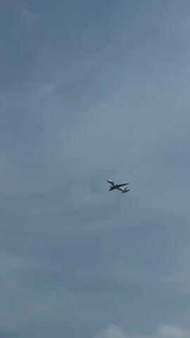 An airplane is flying in the sky. The airplane is white with black and blue markings. It appears to be a commercial jetliner. The sky is overcast with some scattered clouds. The airplane is angled slightly upwards and to the right. The video is shaky, suggesting it was filmed from a moving platform.