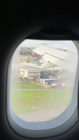 The video shows a view from an airplane window. The airplane wing is visible on the right side of the frame. Below the wing, a runway and grassy area are visible, along with some buildings. The airplane appears to be on the ground, possibly taxiing or preparing for takeoff. The scene is slightly hazy.