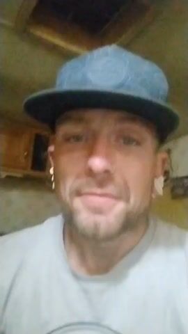 Here is a concise description of the video:

A man with a light beard and mustache wearing a gray t-shirt and a blue and black baseball cap is looking directly at the camera and speaking. He has a nose ring and white earrings. He points toward the camera as he speaks. The background includes a wooden cabinet and ceiling.