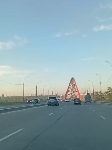The video captures a view from a vehicle driving on a multi-lane highway towards a distinctive red arch bridge. The scene is set during the daytime, with a clear blue sky dotted with sparse white clouds, suggesting fair weather. The sun is casting light from the side, creating some shadows.

The primary subject is the impressive red cable-stayed bridge, which dominates the background with its striking A-frame structure. The bridge's design is characterized by its tall, triangular pylon and the network of cables supporting the roadway. Several vehicles, including cars and a larger truck, are visible on the highway, indicating that this is an active transportation route. The highway itself is well-maintained with clear lane markings.

In the distance, beyond the bridge, a city skyline is faintly visible, suggesting an urban or suburban setting. Streetlights line the sides of the highway, typical of a major road. The overall mood conveyed is one of travel and forward movement, with the bridge serving as a prominent landmark. No discernible text is visible in the video. The location context provided places this scene in Novosibirsk, Russia, which aligns with the architectural style of the bridge.