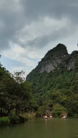 The video shows a river scene with several small boats carrying people. The boats are being paddled through a river that reflects the surrounding lush green vegetation and a towering, tree-covered mountain. The sky is overcast, and the overall atmosphere is one of peaceful exploration and natural beauty.