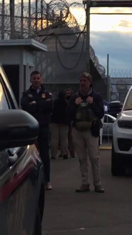 The video shows two men standing outside a building with barbed wire on top of a fence. The man on the left is wearing a dark jacket with the word "ISI" on it, along with a U.S. flag, and the word "AGENT" below the flag. He has his arms crossed. The man in the middle is wearing tan pants, boots, a sweatshirt and a camouflage hat. He also appears to be wearing a vest that says "POLICE SPECIALIST". There are two vehicles, one black and one white, partially visible on either side of the two men. Other people are also visible in the background. The setting appears to be a parking lot outside of a detention facility. The sky is cloudy and it appears to be early morning or late afternoon.