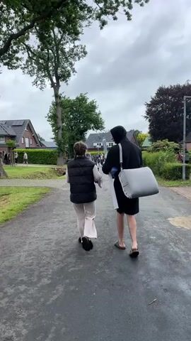 Two individuals walk away from the camera on a paved path. The person on the left is a woman with dark hair, wearing a dark vest over a light-colored long-sleeved top and light-colored wide-leg pants. The person on the right is a man wearing a dark hooded sweatshirt, dark shorts, and sandals. He carries a large, light grey, soft-sided bag over his left shoulder and holds a long, white, rectangular object in his right hand. They walk at a consistent pace. The path is grey and appears to be asphalt. To the left of the path, there is a grassy area, a large tree with green leaves, and several houses in the background. To the right of the path, there is some sparse greenery and more houses in the distance. The sky is overcast.